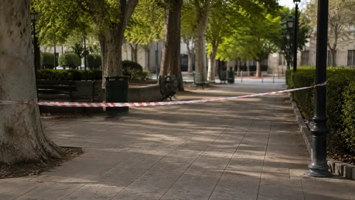 Cinta de seguridad lisa delimitando una vereda vacía en una plaza de barrio con mobiliario urbano, plano medio documental